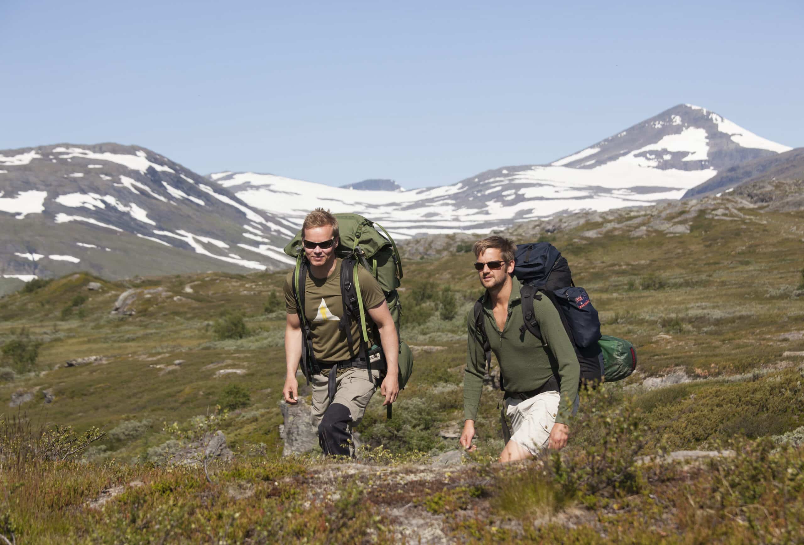 Två män fjällvandrar med snöklädda fjäll i bakgrunden.